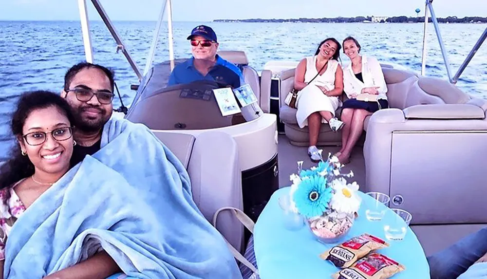 A group of people are enjoying a boat ride with a couple snuggled under a blanket in the foreground and two women sitting with snacks at a table while a man pilots the boat in the background