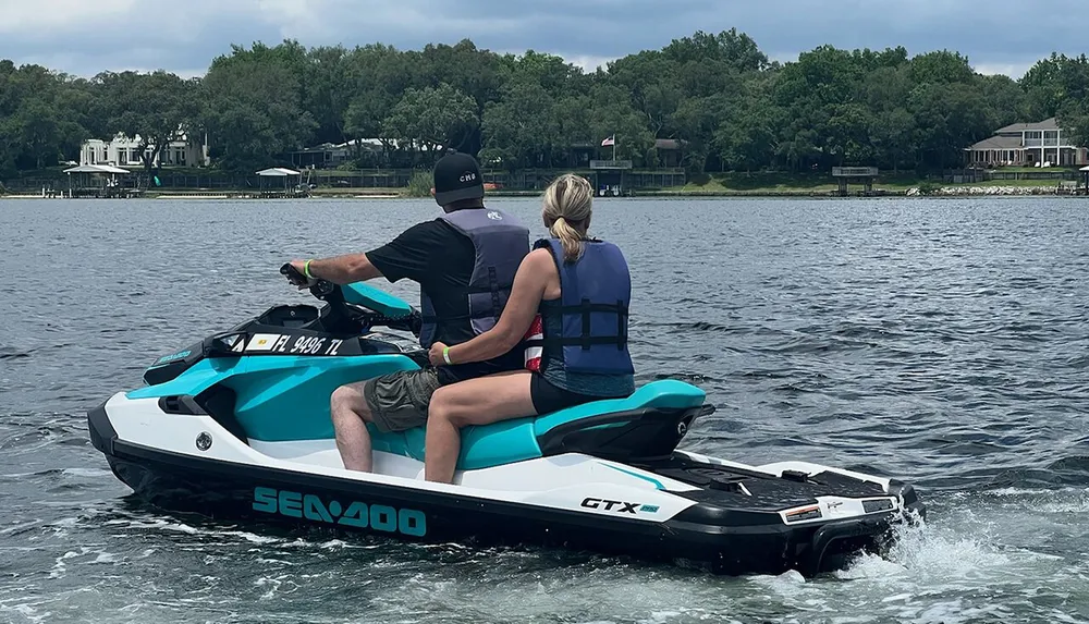 Two people are riding a Sea-Doo personal watercraft on a body of water with lakeside houses in the background