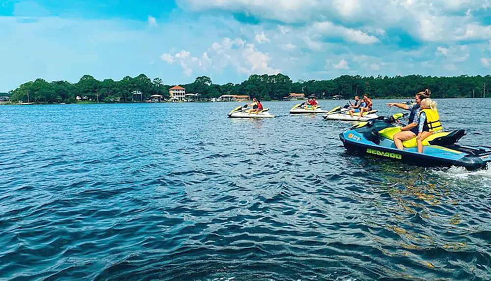 A group of people on personal watercrafts are enjoying a sunny day on the lake with trees and houses in the background