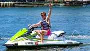 A person is energetically posing with a peace sign while seated on a green and white Yamaha WaveRunner on the water.
