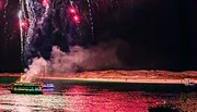 A brightly lit boat sails on a body of water under a night sky, with fireworks exploding above and their reflections shimmering on the water's surface.