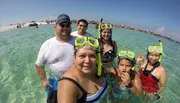 A group of six people wearing snorkeling gear takes a selfie while standing in shallow, clear waters at a beach.