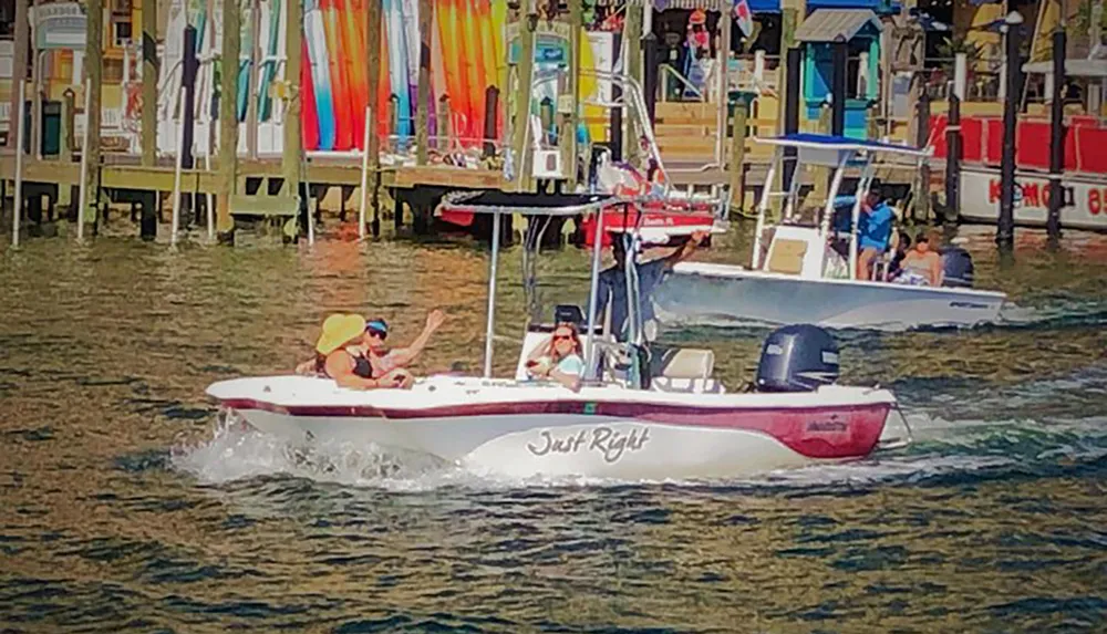 The image shows two people enjoying a sunny day on a small boat named Just Right cruising through the water with colorful paddleboards and docks in the background