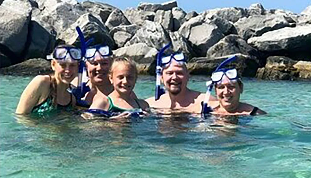 A group of five people is smiling at the camera while wearing snorkeling gear in clear shallow water with rocks in the background