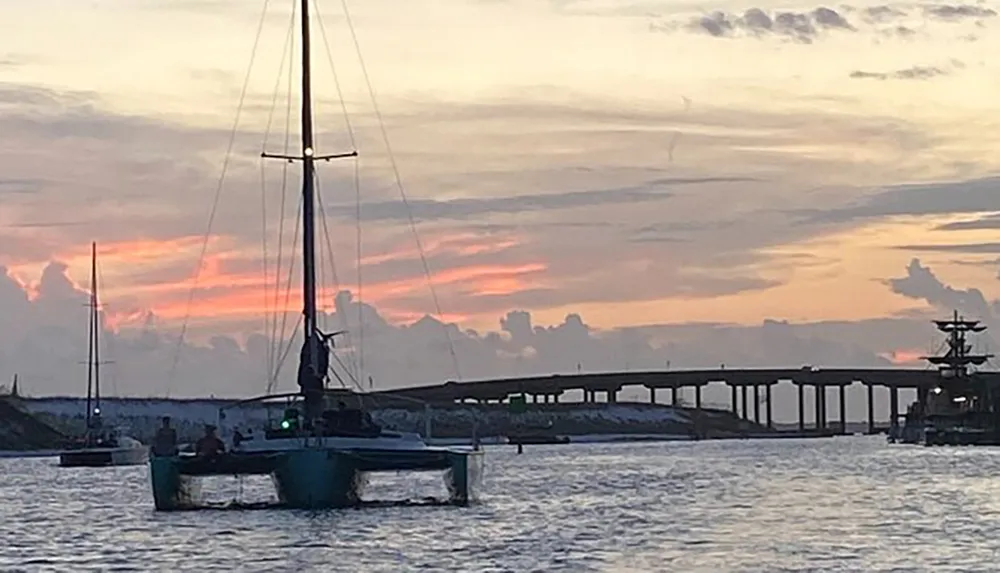 A sailboat is silhouetted against a vibrant sunset with a bridge in the background and other boats on the water