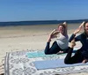 Two people are smiling and posing in a yoga position on a beach towel by the ocean
