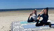 Two people are smiling and posing in a yoga position on a beach towel by the ocean.