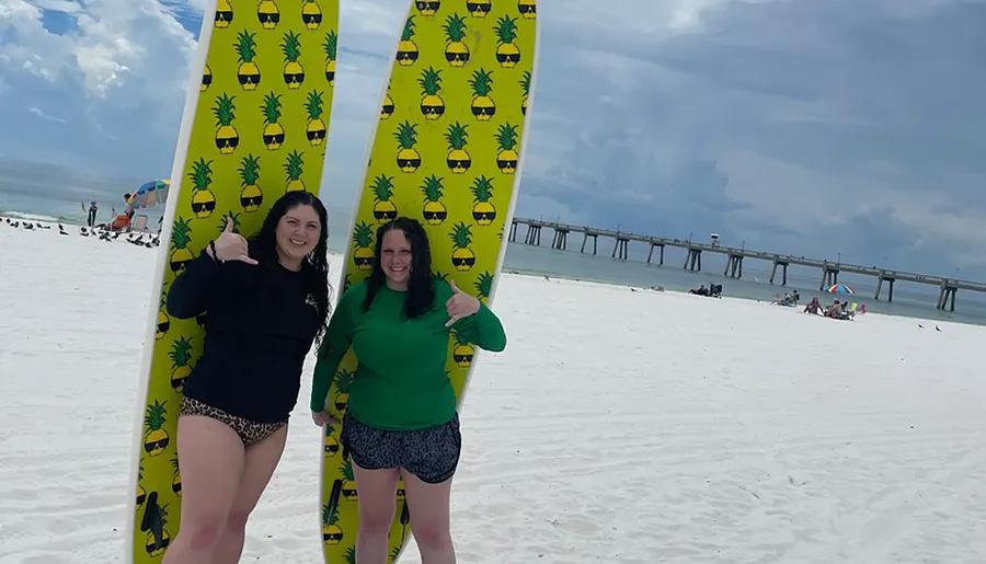 Two people are posing with bright yellow surfboards decorated with pineapple patterns on a white sandy beach with a pier in the background.