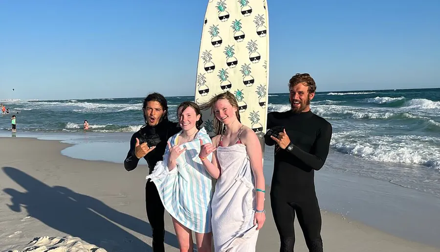 Three people wrapped in beach towels pose with two smiling surfers holding a surfboard on a sunny beach.