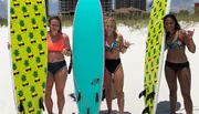 Three smiling people are standing on a sunny beach, each with a surfboard, ready to enjoy surfing.
