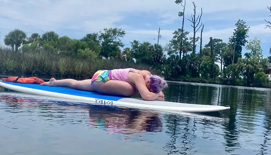 A person with purple hair is lying face down on a paddleboard in calm waters, appearing to be resting or sunbathing.