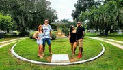 Four people are posing for a photo in a green park with a fountain and Spanish moss-draped trees in the background.