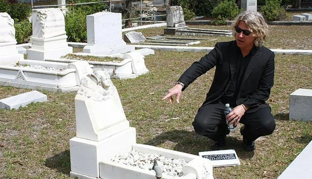 A person is crouching beside a damaged statue on a grave gesturing with their hand presumably discussing or explaining the situation