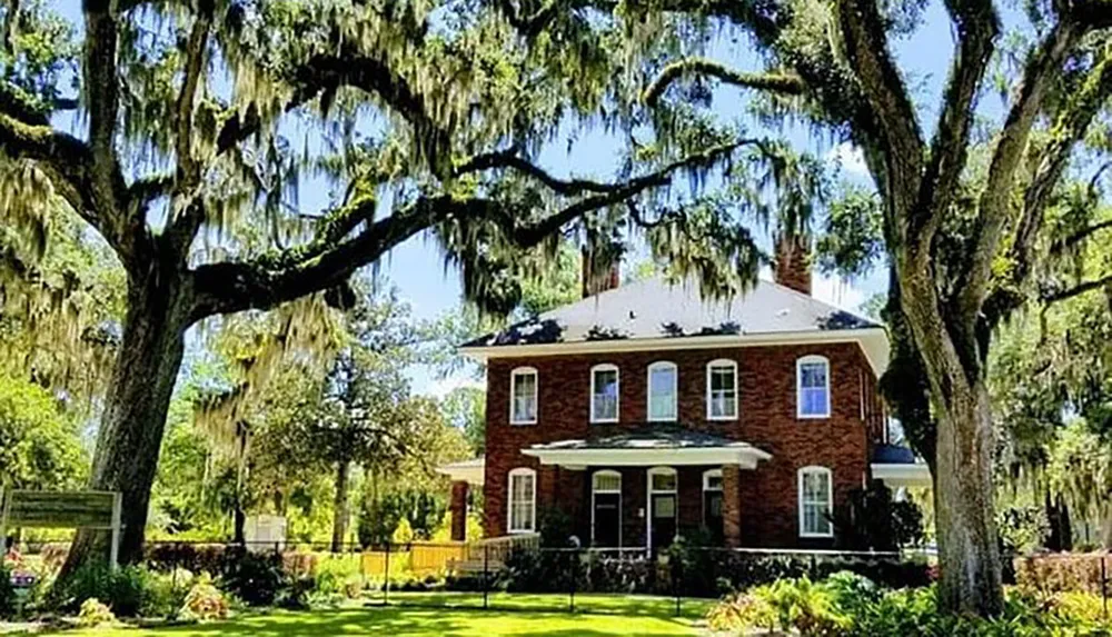 A stately brick house framed by majestic trees draped with Spanish moss under a sunny sky