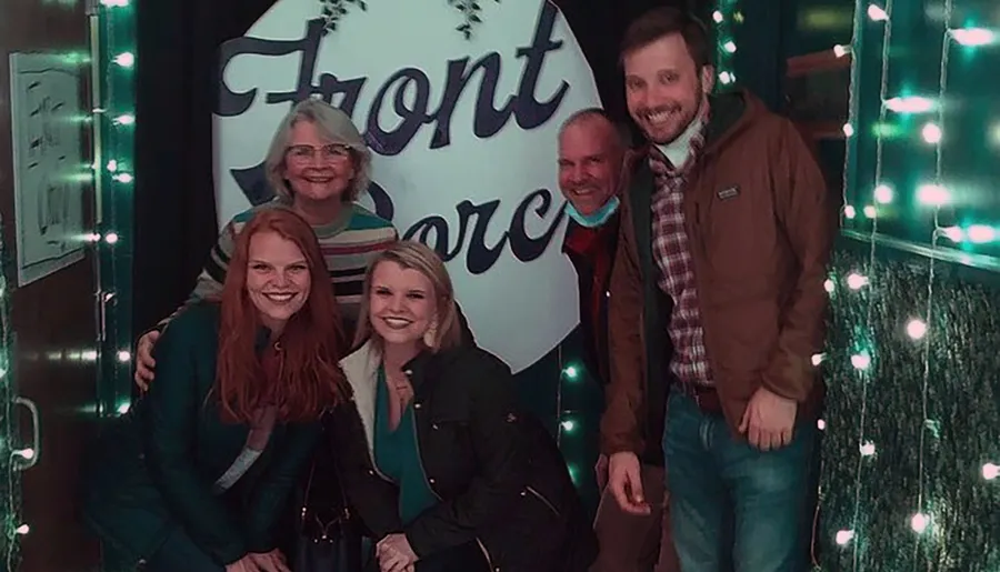 A group of five people are smiling for a photo in front of a sign that reads Front Porch decorated with twinkling lights.