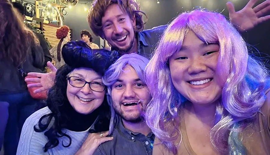 Four cheerful people are posing for a selfie, with two of them wearing colorful wigs, seemingly enjoying a festive or costume event.