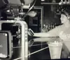 A black and white photo shows a woman wearing headphones behind a bar being filmed by a camera with a viewfinder displaying her image