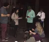 A group of people are gathered outdoors at night some standing and some squatting looking at or using their smartphones