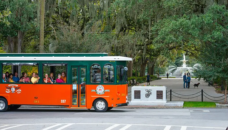 A colorful trolley full of passengers travels near a park with a fountain and Spanish moss-draped trees in the background.