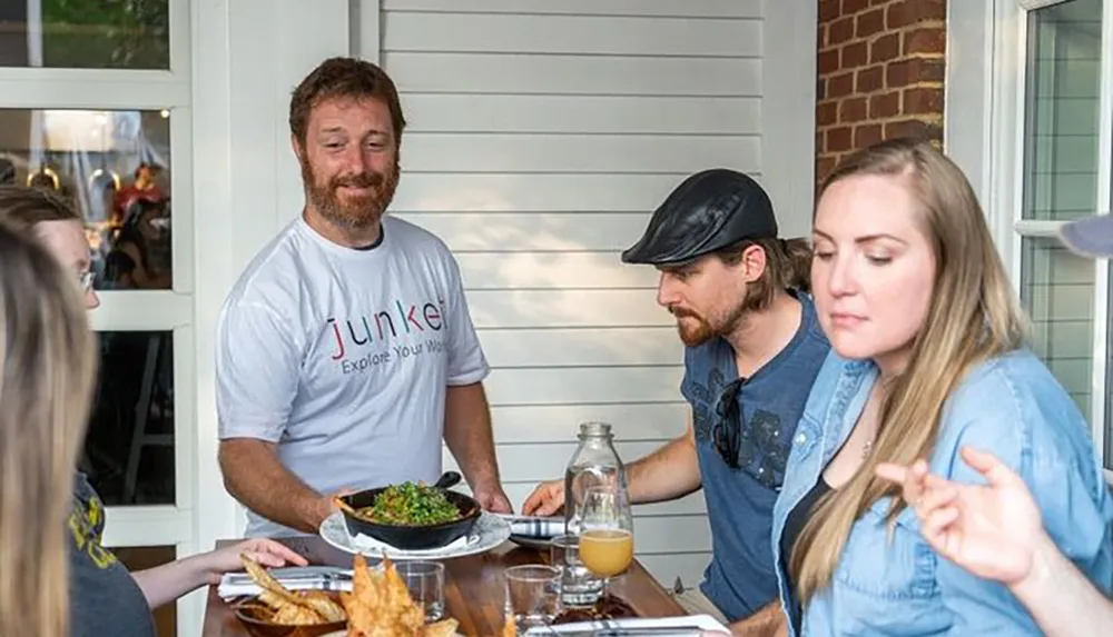 A group of people are enjoying a meal together at an outdoor dining setting with one person smiling at the camera while the others are engaged in conversation or looking at their food