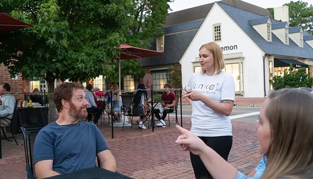 A woman is speaking to seated people at an outdoor restaurant patio with umbrellas and diners in the background