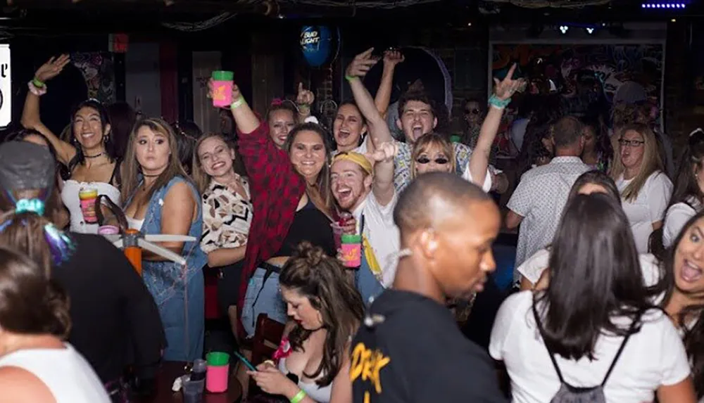 A lively crowd of people with raised hands and drinks is enjoying themselves at what appears to be an indoor night club or party venue