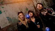 Three individuals appear to be engaged in a ghost hunting activity, displaying expressions of surprise and curiosity, in a dilapidated interior environment.