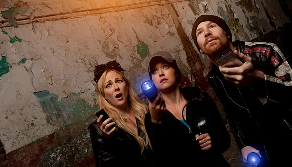 Three individuals appear to be engaged in a ghost hunting activity displaying expressions of surprise and curiosity in a dilapidated interior environment