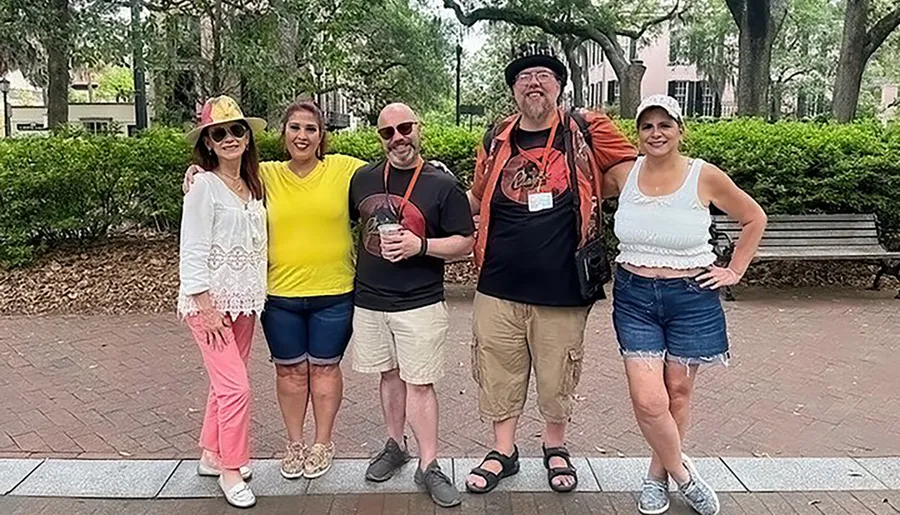 Five people are posing for a group photo outdoors, smiling and dressed in casual attire.