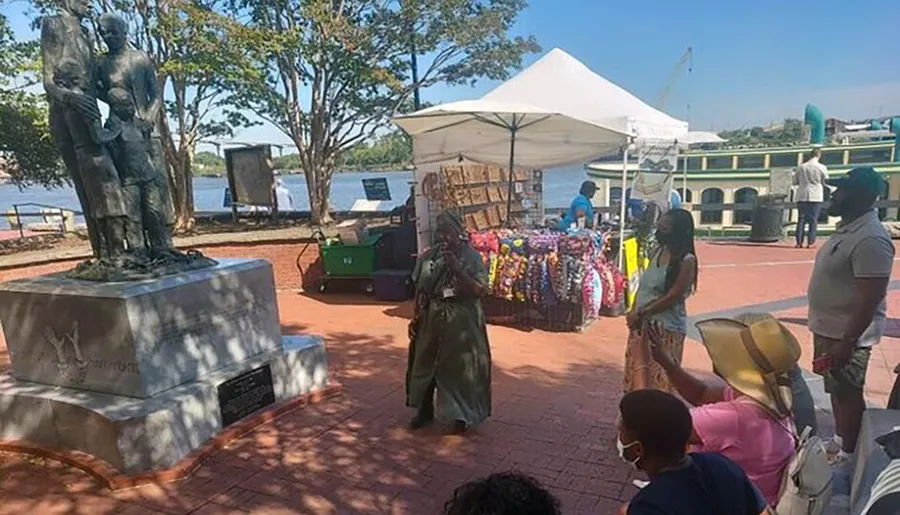 A statue of a group of individuals stands in a park area near a waterfront, while people nearby engage in various activities like browsing a vendor's tent, taking a photo, and walking.