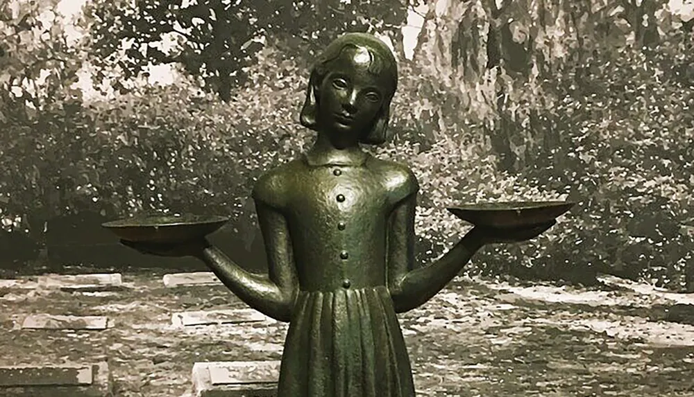 The image shows a bronze statue of a girl with outstretched arms holding two bowls set against a backdrop of trees and foliage