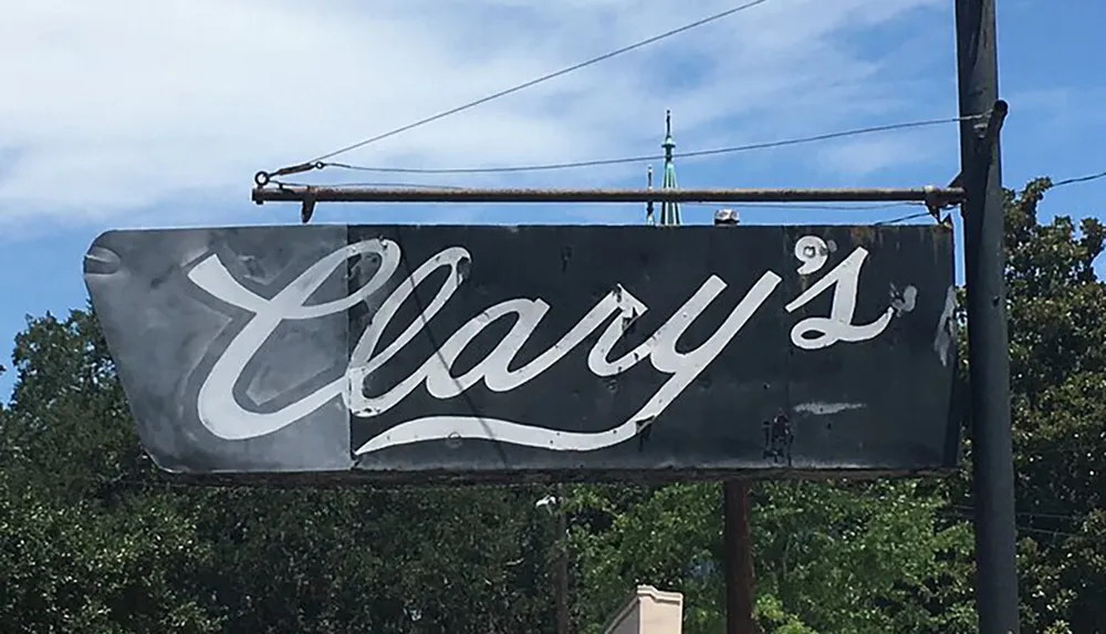 The image shows a worn-out and faded sign with the word Clarys on it suspended by a metal frame against a backdrop of clear blue sky and trees