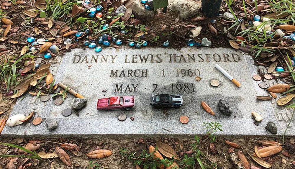 The image shows a memorial stone for Danny Lewis Hansford adorned with a variety of small items and trinkets set amidst a ground covered with leaves