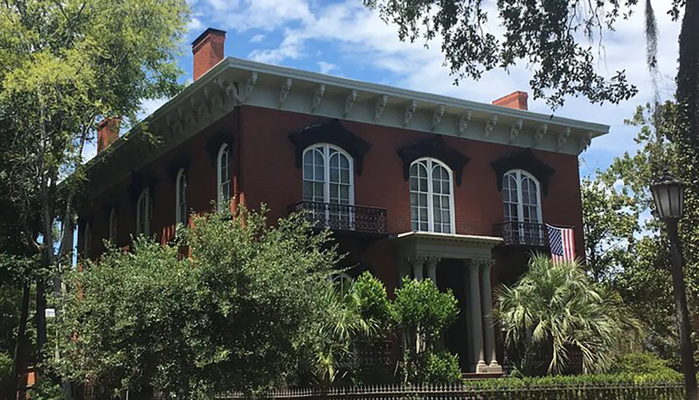 The image displays a stately red brick house with white columns arched windows and an American flag hanging from the second floor all surrounded by lush greenery and trees