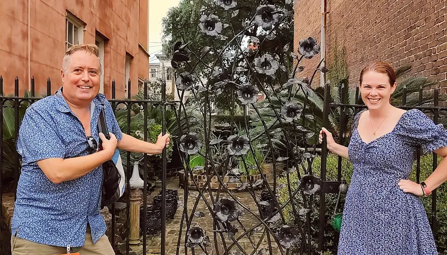 A man and a woman are smiling and posing next to a decorative metal gate adorned with flower designs in an outdoor setting.