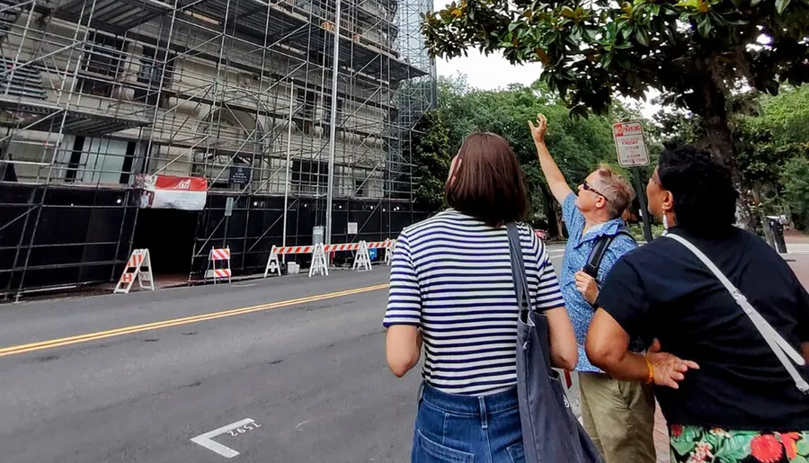 Three individuals are observing a construction site with scaffolding while standing on a city sidewalk.