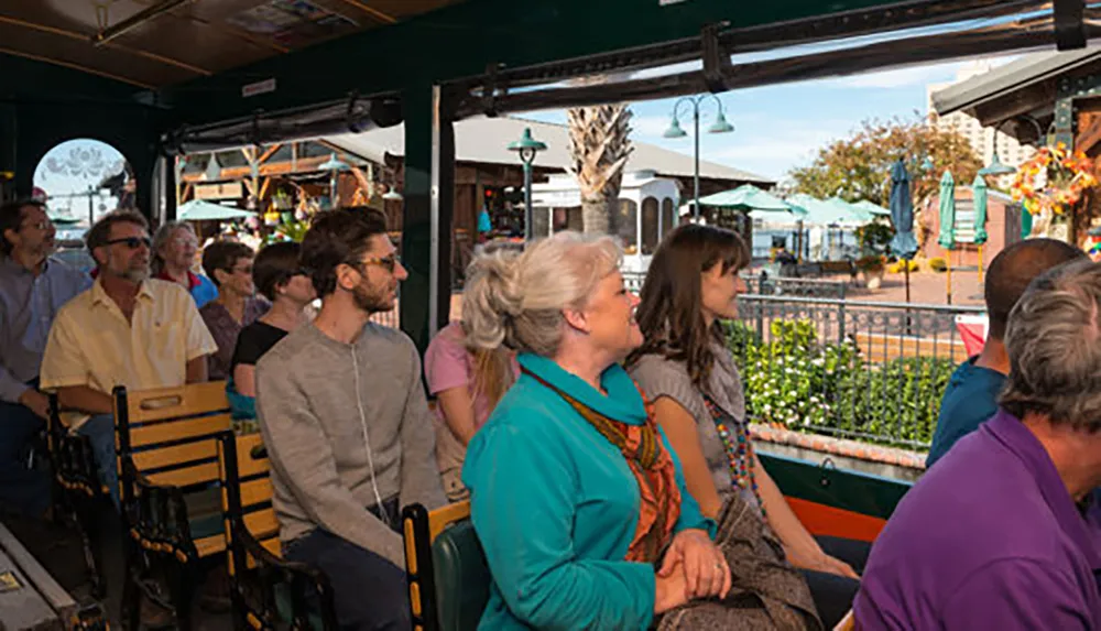 A group of people is sitting inside a tram or open vehicle looking attentively to their right at something outside the frame in what appears to be a vibrant outdoor setting