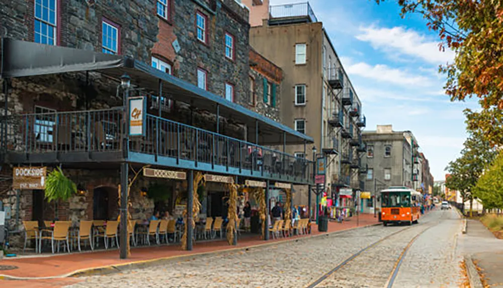 An orange trolley travels along a cobblestone street by old buildings with a second-story balcony restaurant in what appears to be a historic urban area