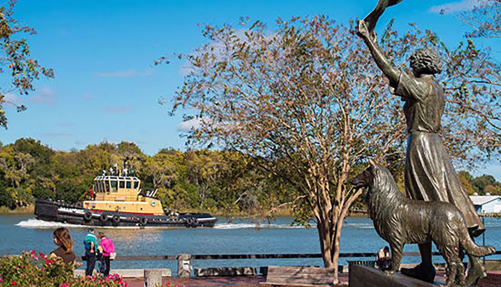 A statue of a woman with her arm raised next to a dog overlooks a river where a tugboat is passing by with onlookers enjoying the serene scene