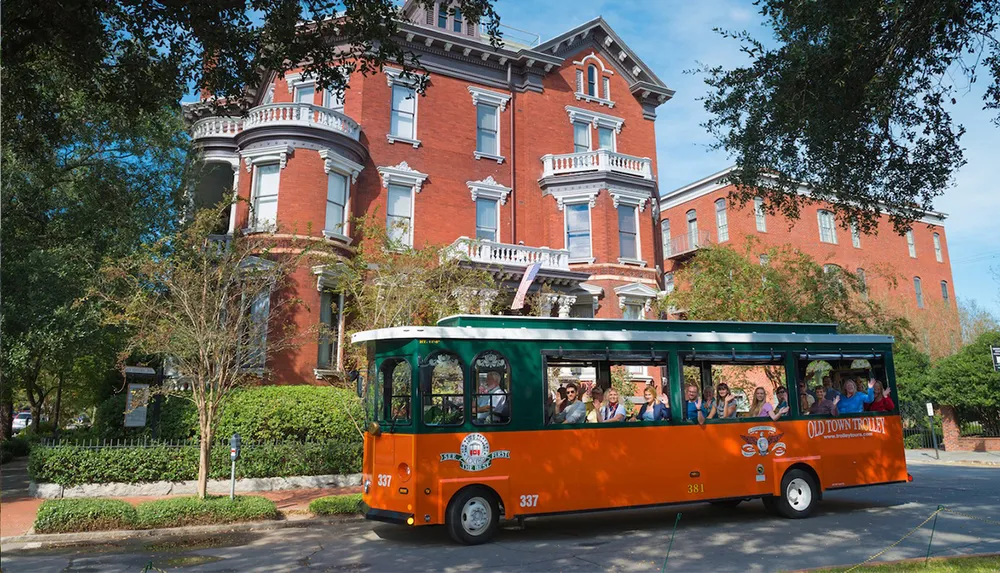 An orange and green trolley filled with passengers tours by a stately red brick Victorian-style building on a sunny day