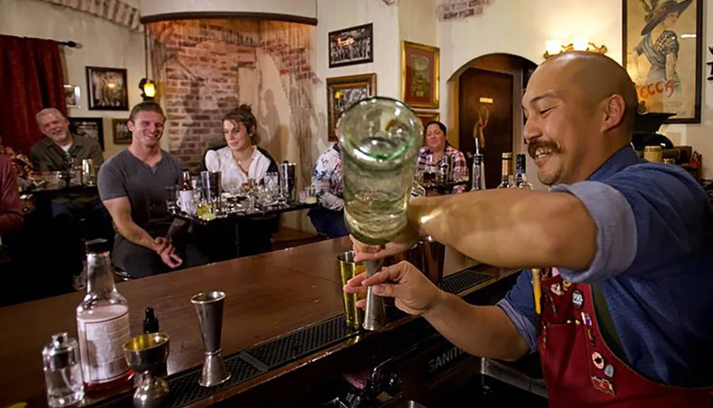 A bartender is performing a bottle trick in front of amused patrons at a warmly lit traditional-style bar