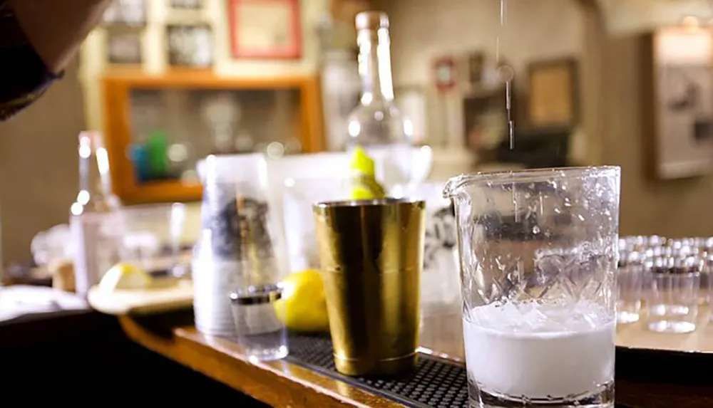 A cocktail preparation scene with a focus on a glass of frothy liquid likely a shaken drink on a bar with various bartending tools and ingredients in the background