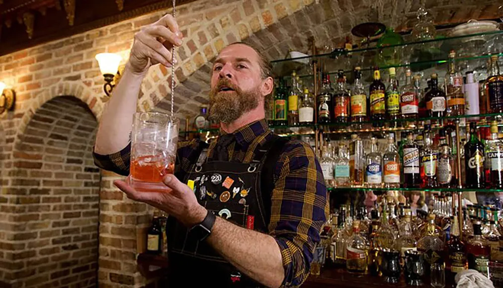 A bearded bartender carefully stirs a cocktail in a glass tumbler against the backdrop of a well-stocked bar