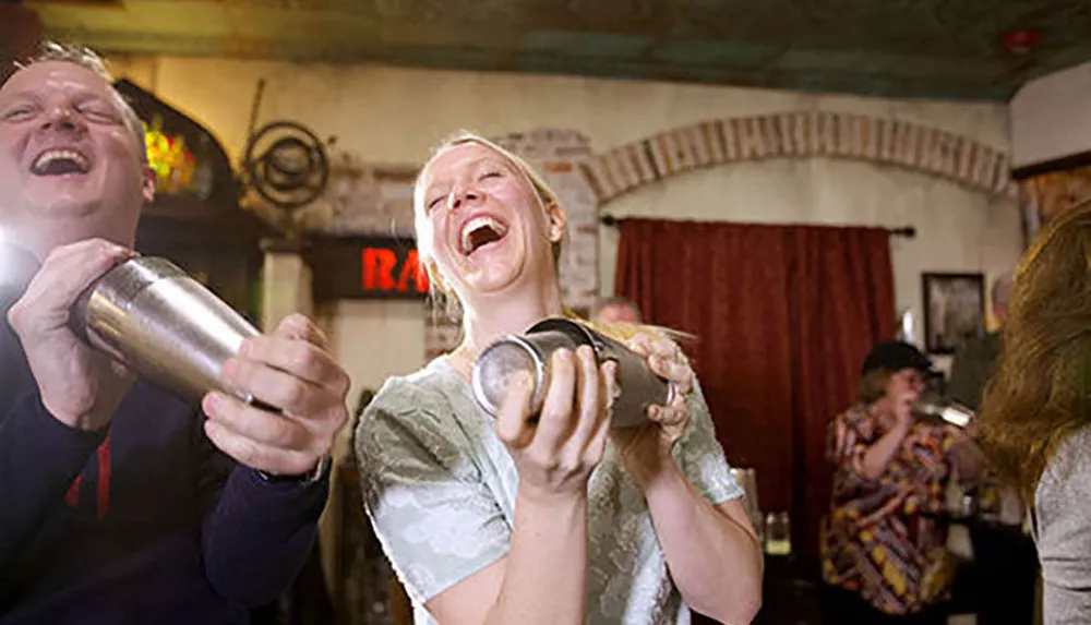 Two people are laughing heartily while shaking cocktail shakers inside a warmly lit room that appears to be a bar or restaurant with others enjoying themselves in the background