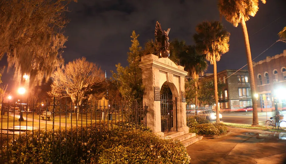 An ornate gate with an eagle sculpture atop surrounded by night-lit trees and street lights stands as an entryway or monument in an urban evening setting