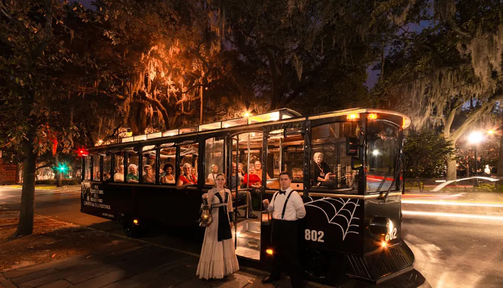 A trolley bus with passengers is parked at night under tree-lined streets accompanied by two individuals in period clothing by its side