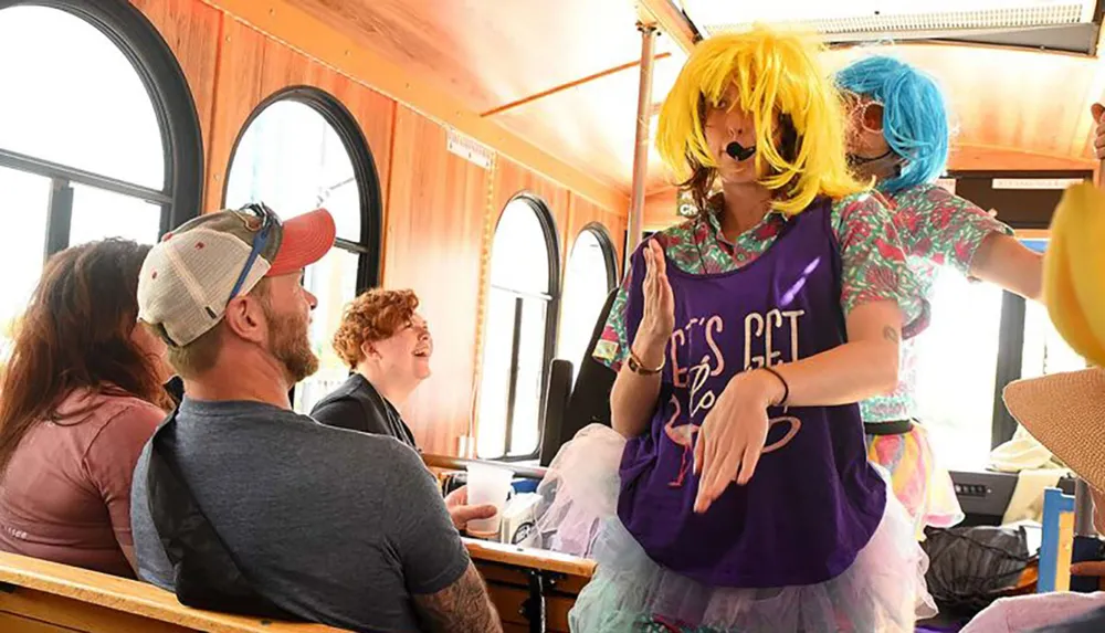 Two performers in colorful and whimsical costumes are entertaining smiling passengers aboard a sunlit train car