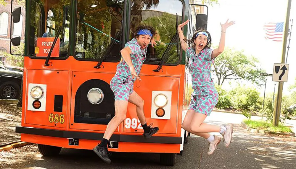 Two people in matching outfits appear to be joyfully jumping off the side of a bright orange trolley or tour bus