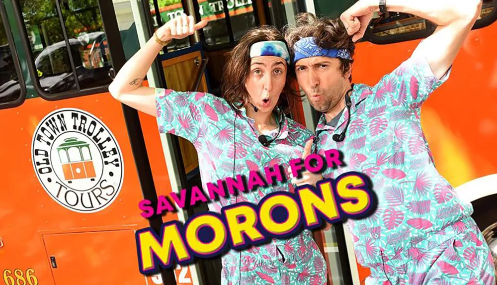 Two individuals wearing matching tropical-themed clothing and headbands pose humorously in front of a trolley promoting a tour named Savannah for Morons