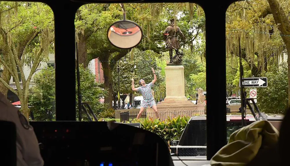 A person is posing with arms outstretched in a spirited manner before a statue as seen through the front windshield of a vehicle with a glimpse of the drivers environment